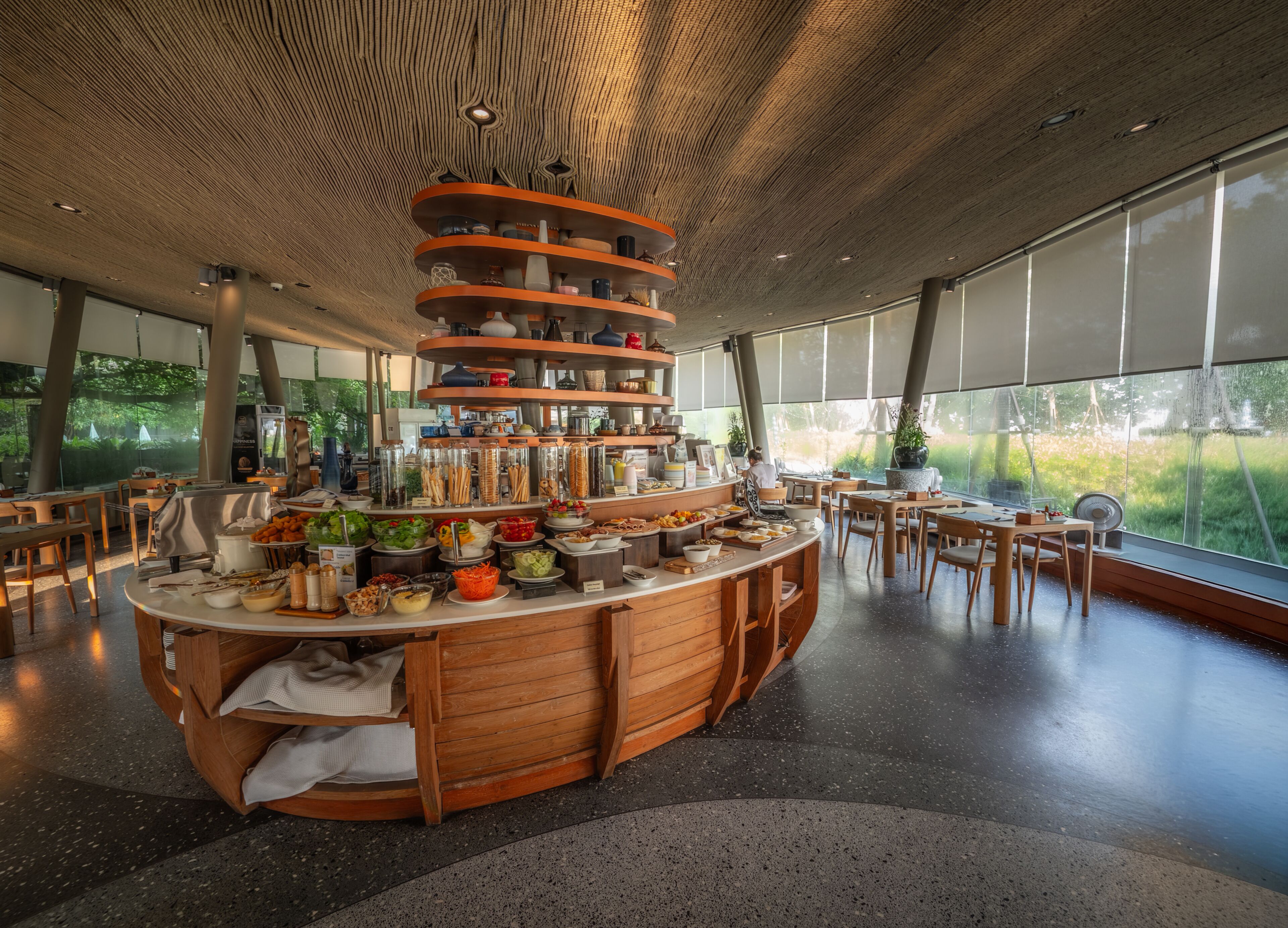 Un restaurante moderno con una barra de madera central, diversas estaciones de comida y grandes ventanales con vistas a zonas verdes.