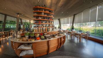 Un restaurante moderno con una barra de madera central, diversas estaciones de comida y grandes ventanales con vistas a zonas verdes.
