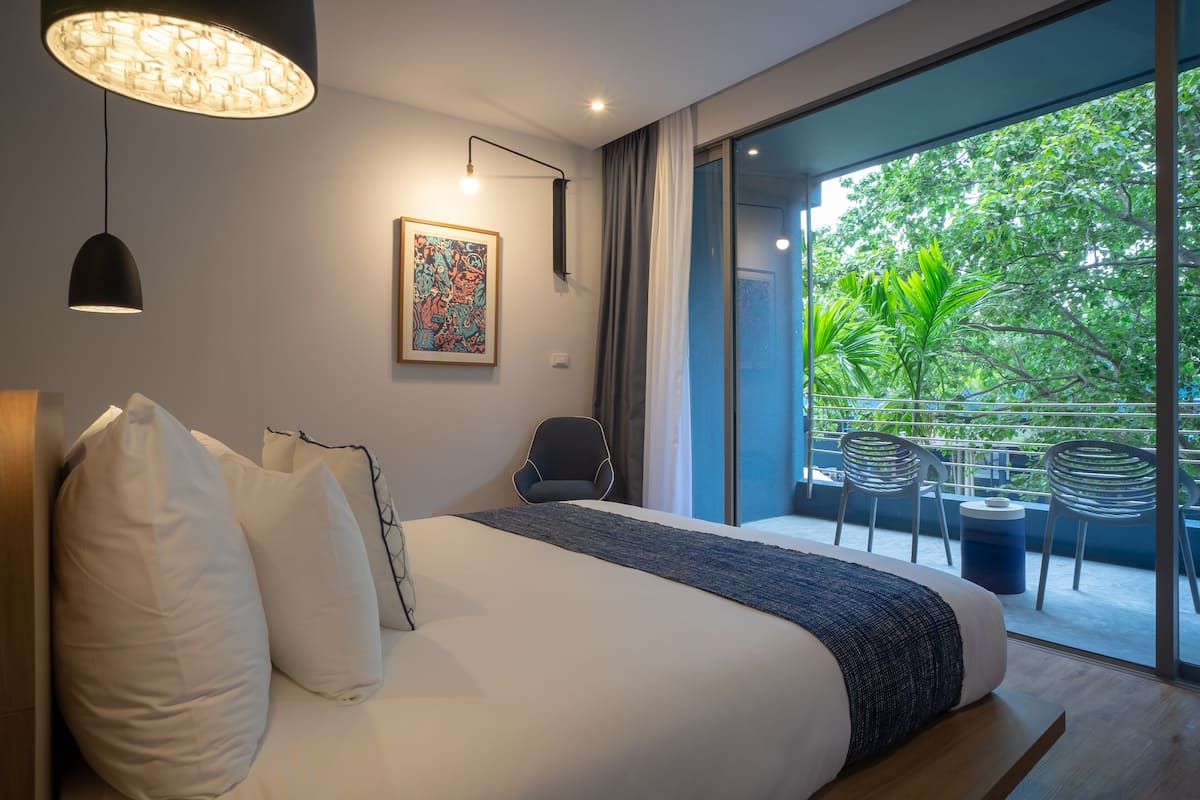 A modern hotel room with a large bed, a view of the outdoors, and a dining area with chairs.