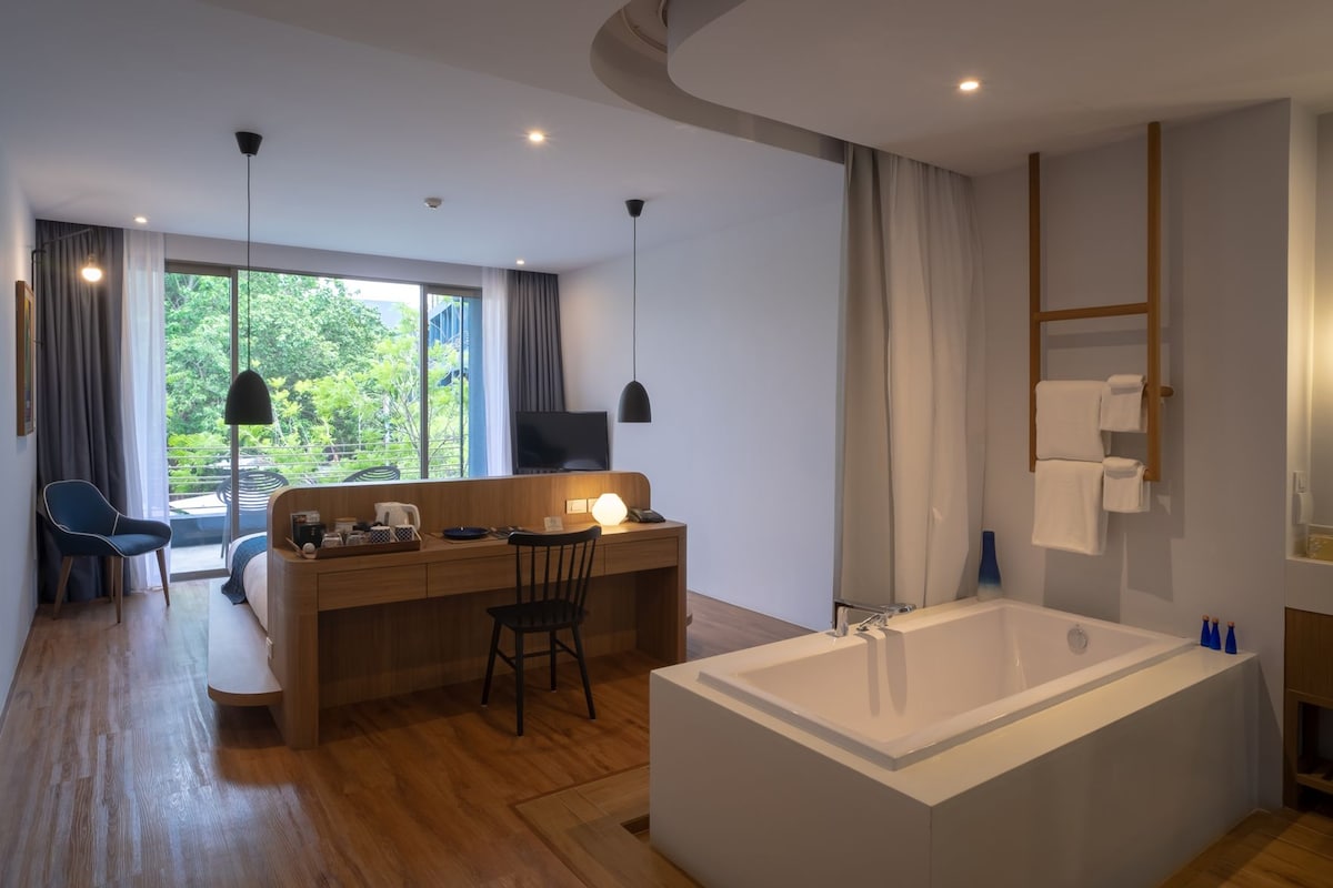 A modern hotel room with a wooden desk, a large bathtub, and a balcony with a view of greenery.