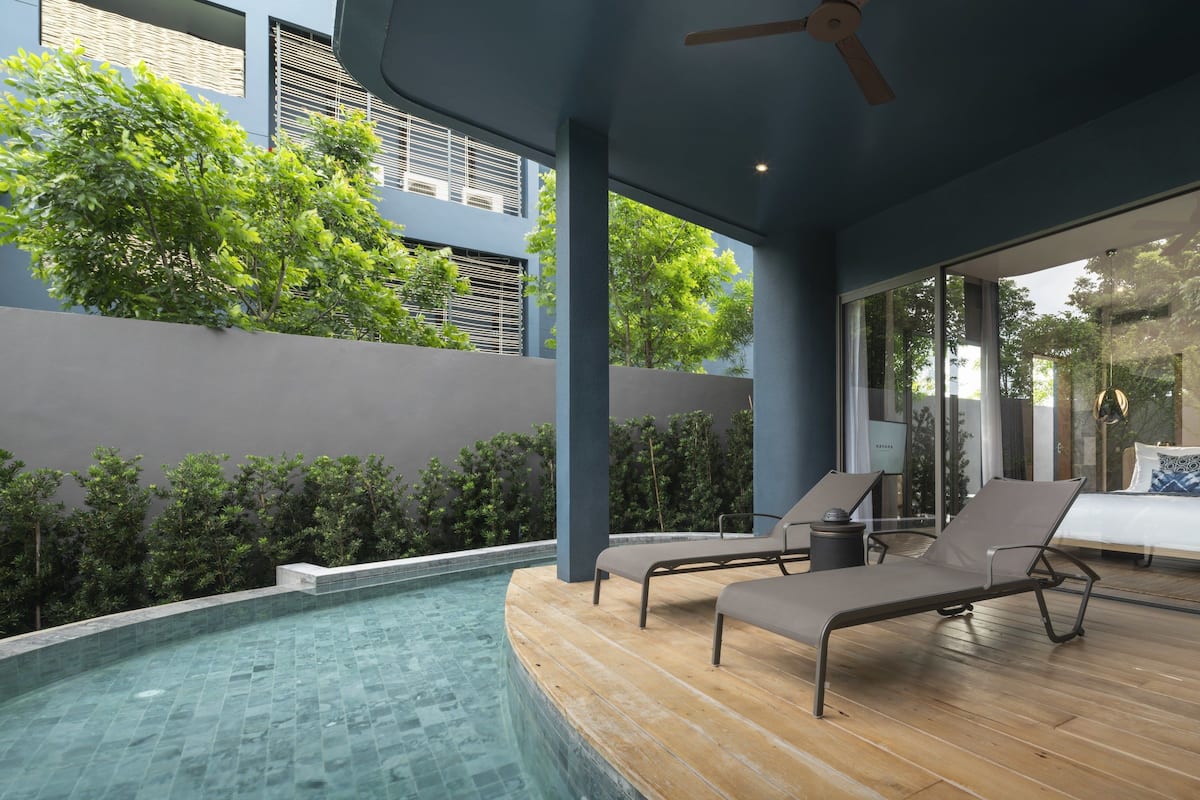 A modern outdoor pool area with lounge chairs, a ceiling fan, and a view of a building with large windows.