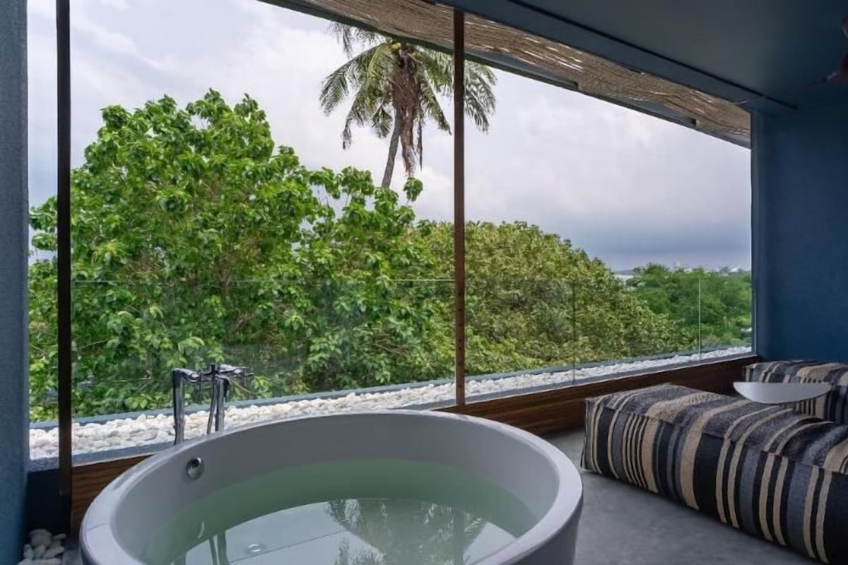 A modern bathroom with a large bathtub, a seating area with striped cushions, and a view of a tropical landscape.