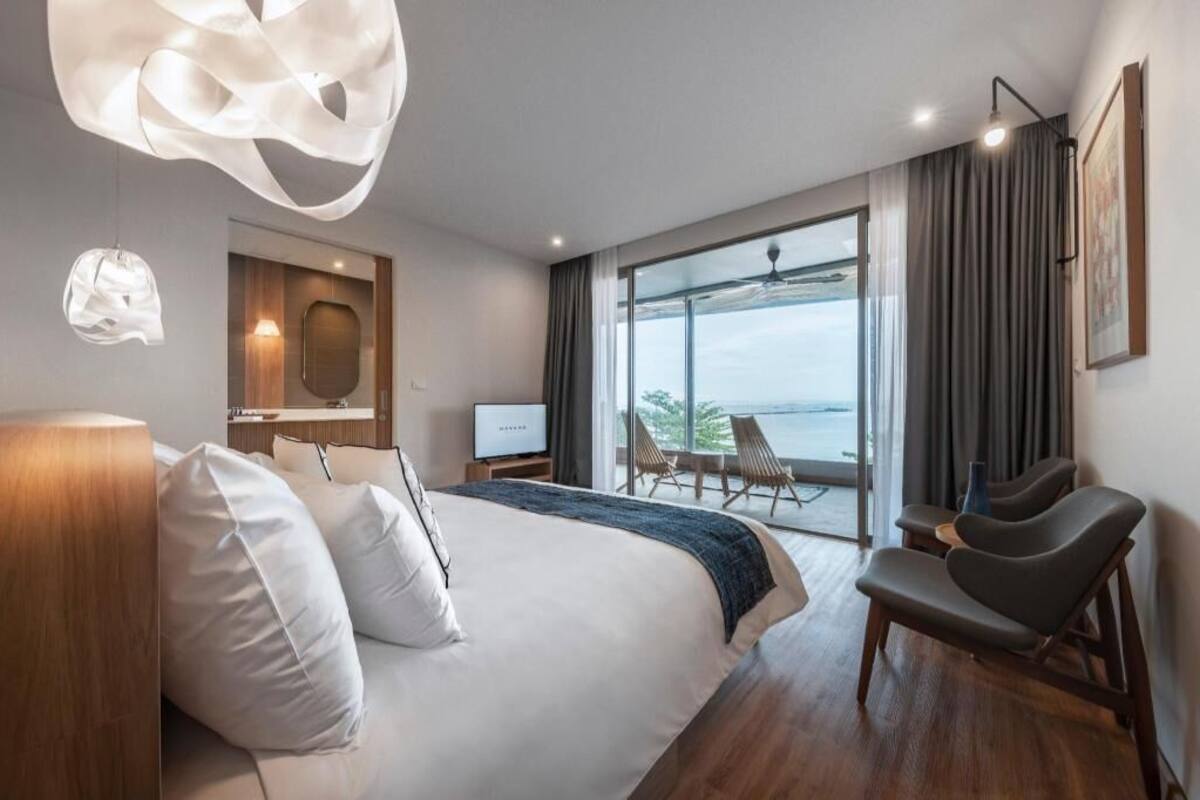 A modern hotel room with a large bed, a desk, a laptop, a chair, and a view of the sea.