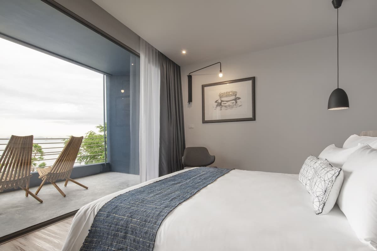 A modern hotel room with a large bed, a balcony with two wooden chairs, and a framed picture on the wall.