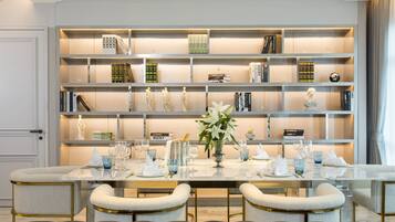 Un área de comedor con una mesa larga dispuesta para cuatro personas, flanqueada por sillas acolchadas, una estantería con libros y objetos decorativos, y un jarrón con flores sobre la mesa.