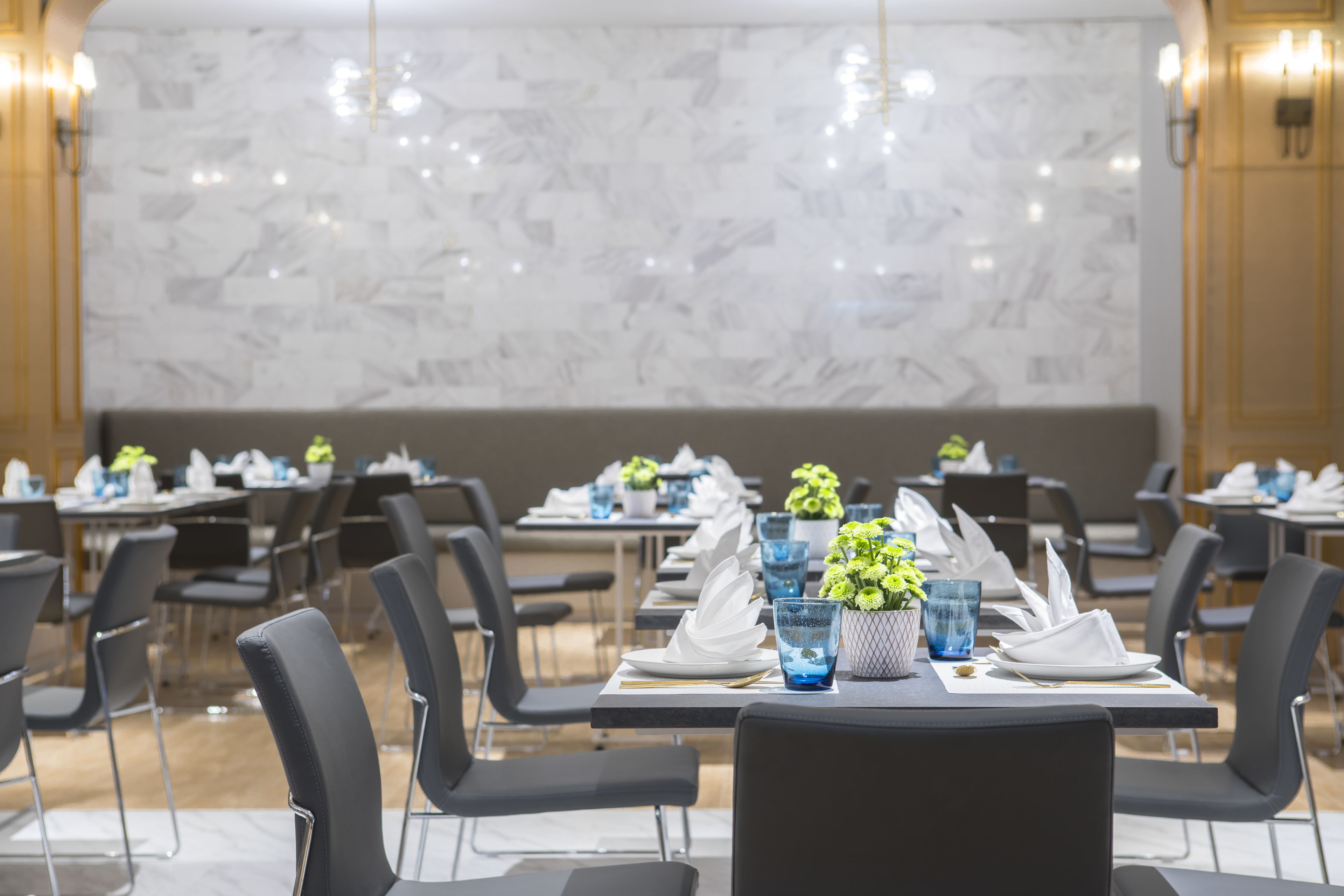 A dining area with marble walls, black chairs, and tables set with white plates, blue glasses, and green centerpieces.
