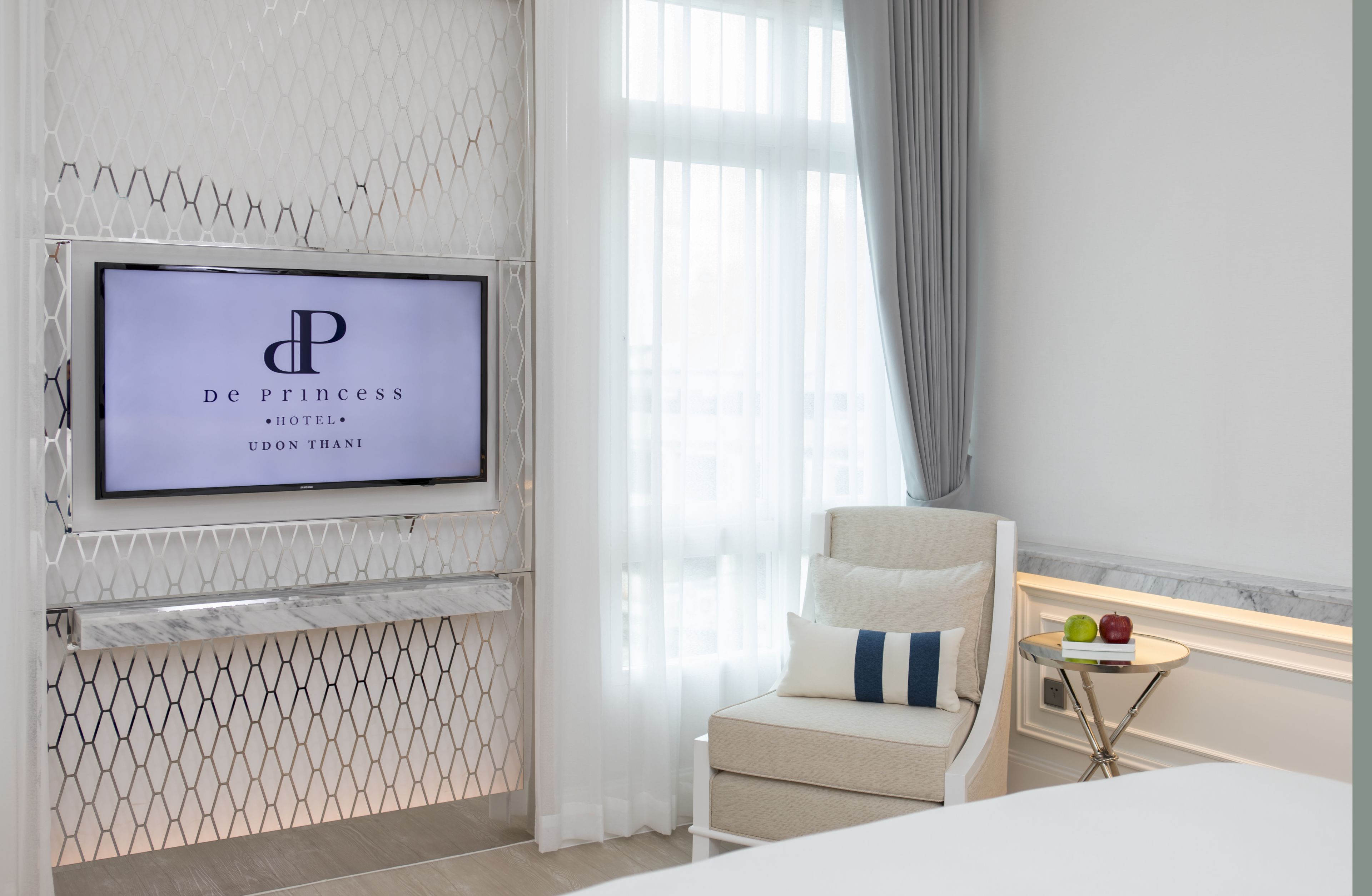 A modern hotel room with a TV displaying the hotel name, a comfortable sofa, a small table with apples and books, and a view of the outside through large windows.