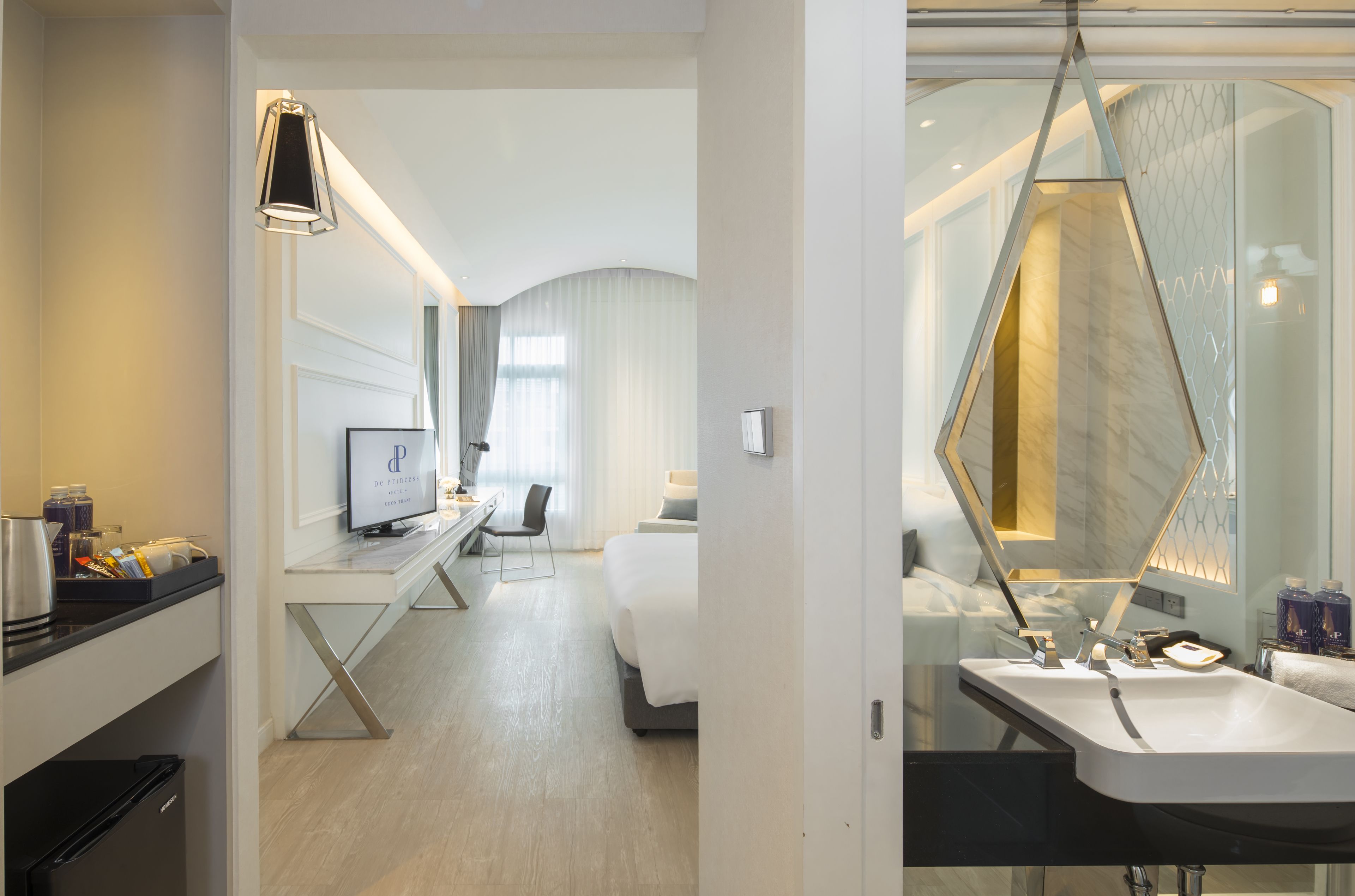 A modern hotel room with a desk, computer, bed, and a bathroom with a large mirror and sink.