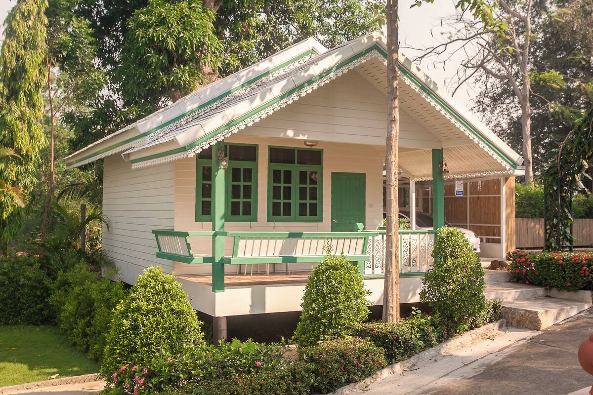 Una casa pequeña de un solo piso con un porche y contraventanas verdes, rodeada de jardines cuidados.