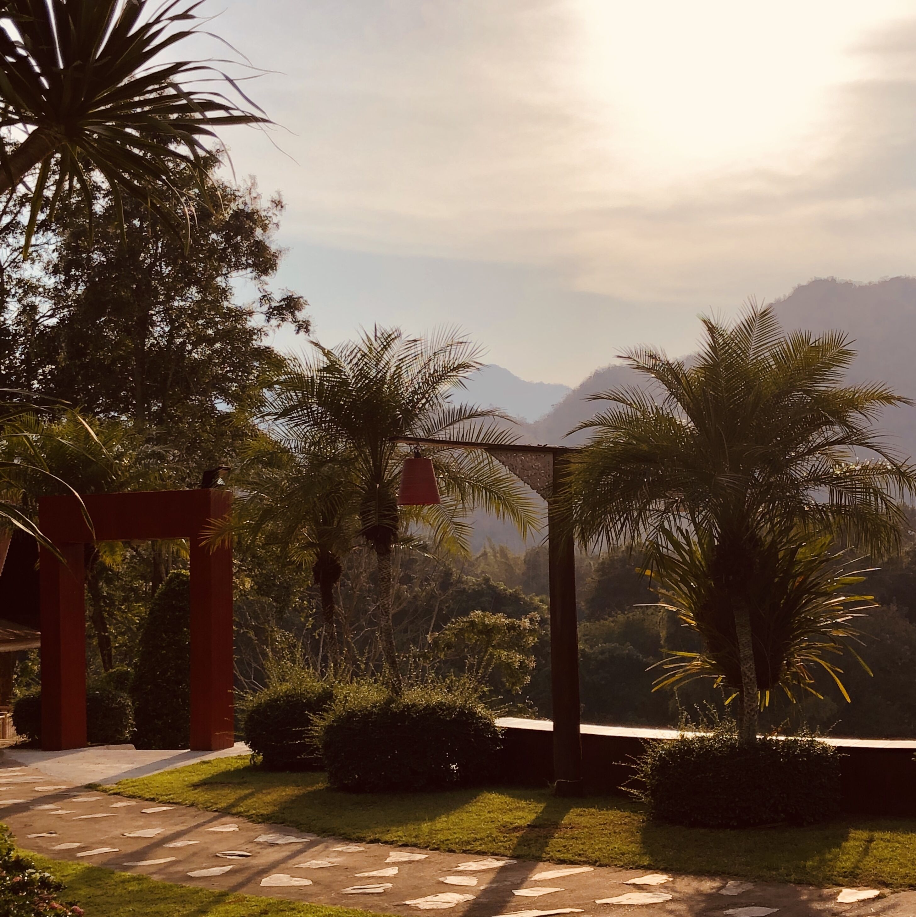 A pathway leading to a red archway surrounded by lush greenery and palm trees, with a mountainous backdrop.