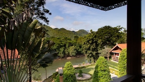 A view from a balcony overlooking a river, lush greenery, and a traditional building with a red roof. - Baanpufa Resort (Sai Yok)