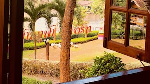 A view through an open window of a lush garden with a tall tree, a palm, and a water body. - Baanpufa Resort (Sai Yok)
