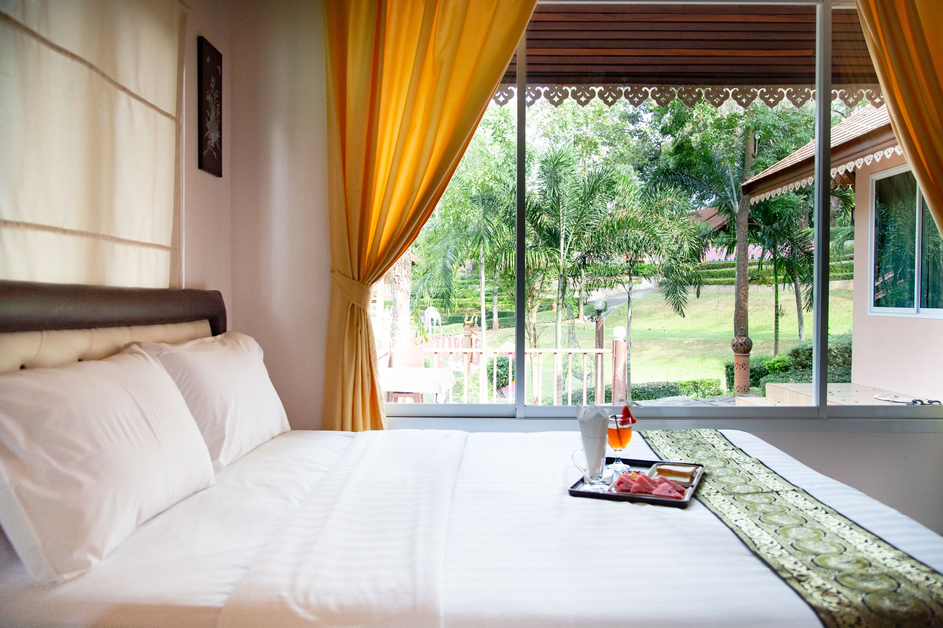 A hotel room with a bed, a tray with food and drink, and a view of a garden and building outside.
