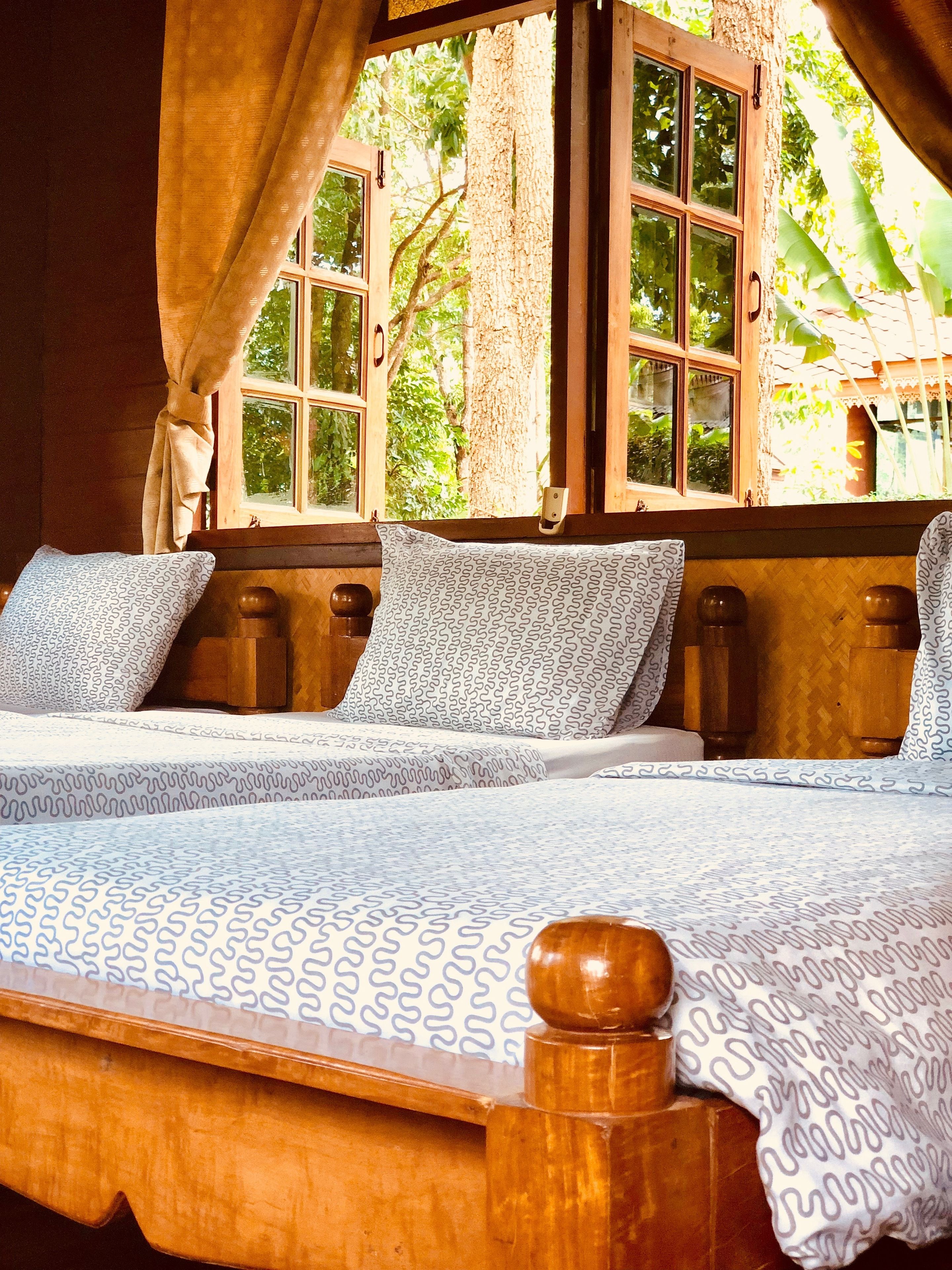 Ein hölzernes Bett mit gemusterten Betttextilien, ein Fenster mit Vorhängen und ein Blick auf Grün außerhalb.