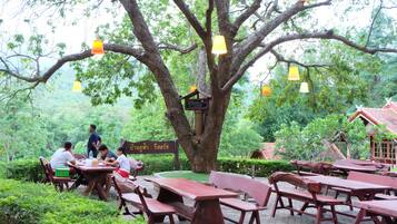 An outdoor dining area with wooden tables and benches under a large tree, surrounded by greenery.