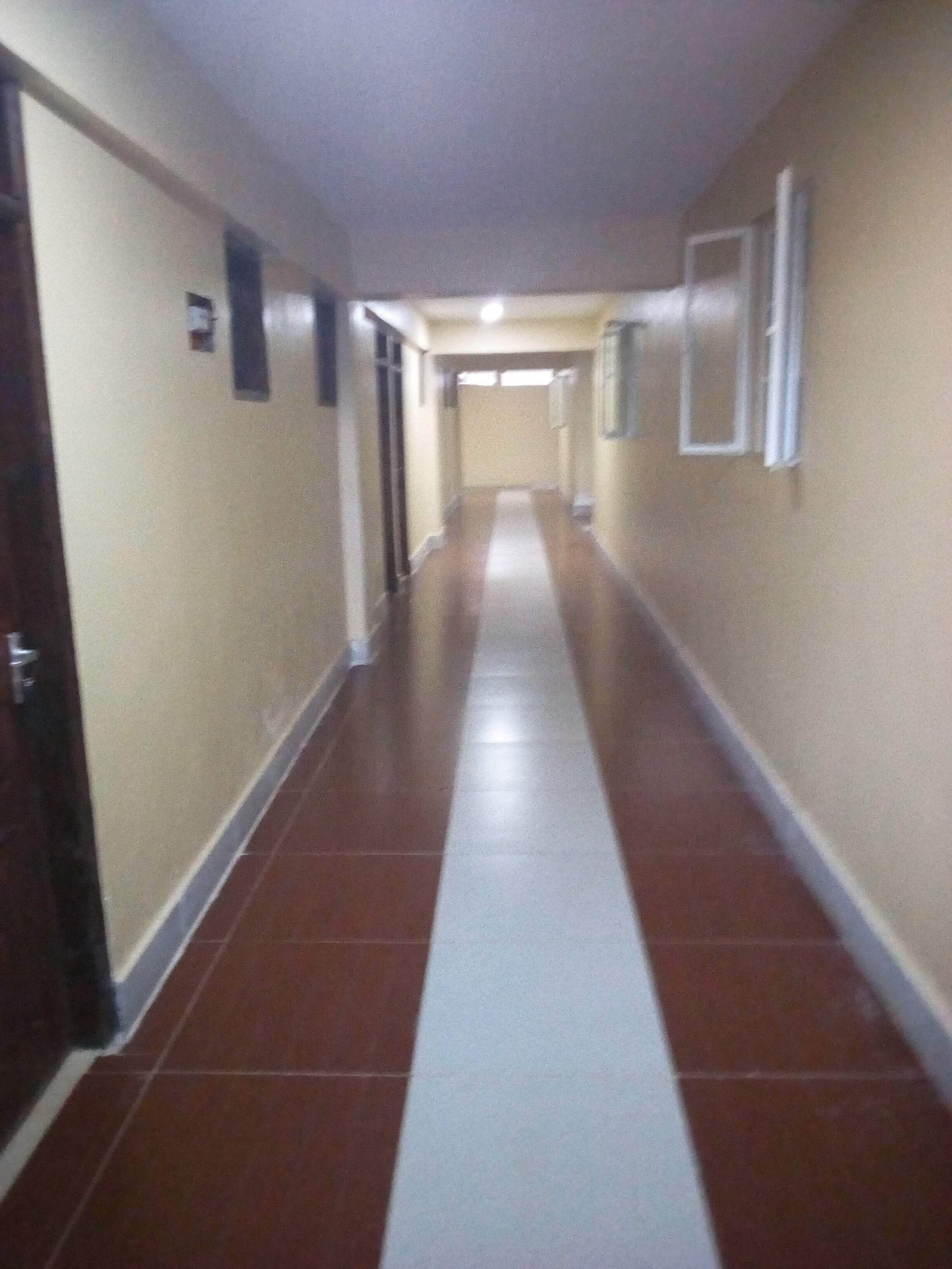 A long, narrow hallway with a red and white striped floor, doors on both sides, and a plain wall with a small sign on the left.