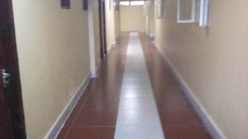 A long, narrow hallway with a red and white striped floor, doors on both sides, and a plain wall with a small sign on the left.