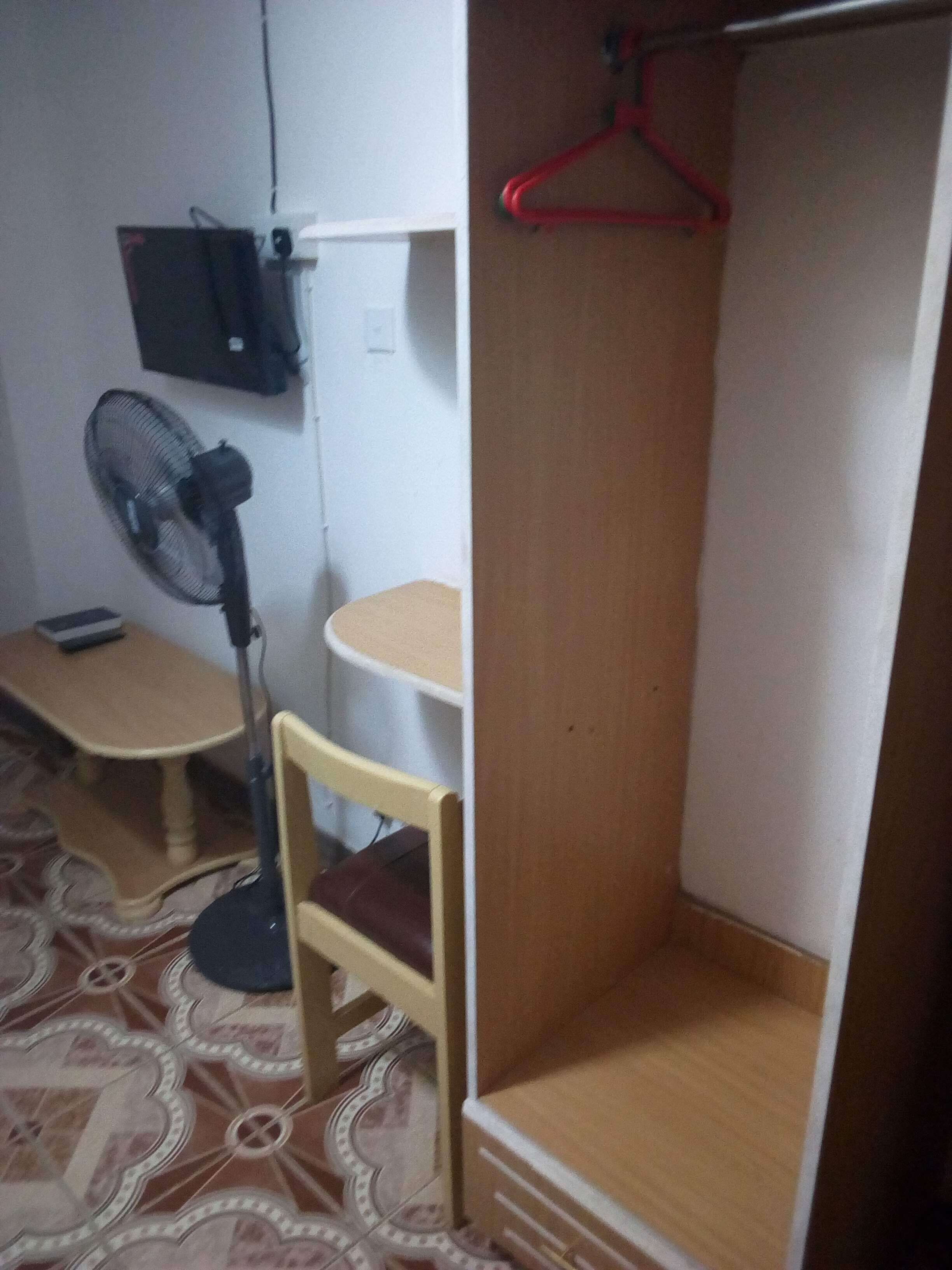 Una habitación compacta con un armario de madera, una mesita pequeña, una silla, un escritorio, un perchero rojo colgante y un ventilador de pie.