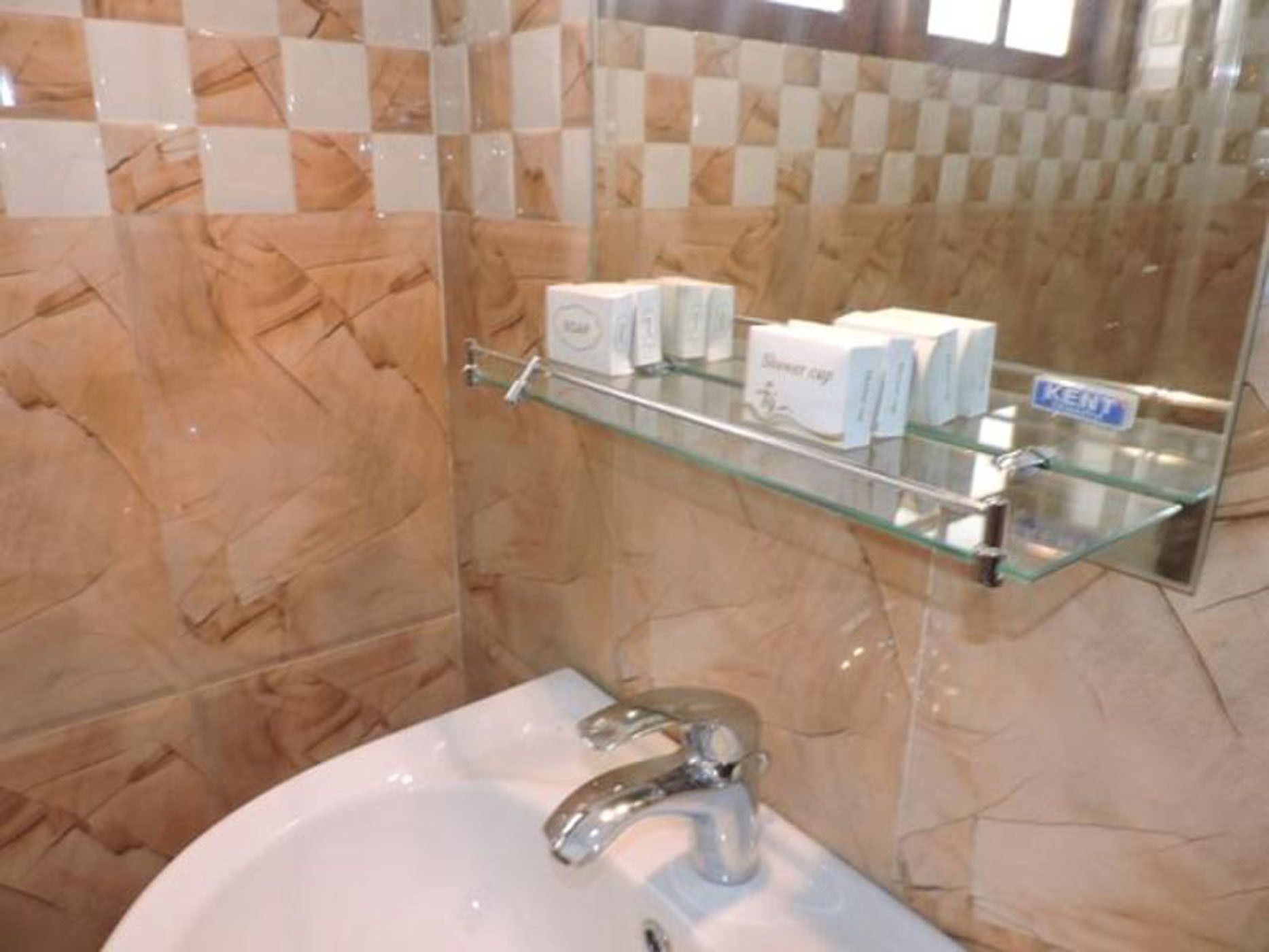 Une salle de bain avec un mur en marbre, une étagère en verre où sont rangés les articles de toilette, et un lavabo blanc avec un robinet chromé.