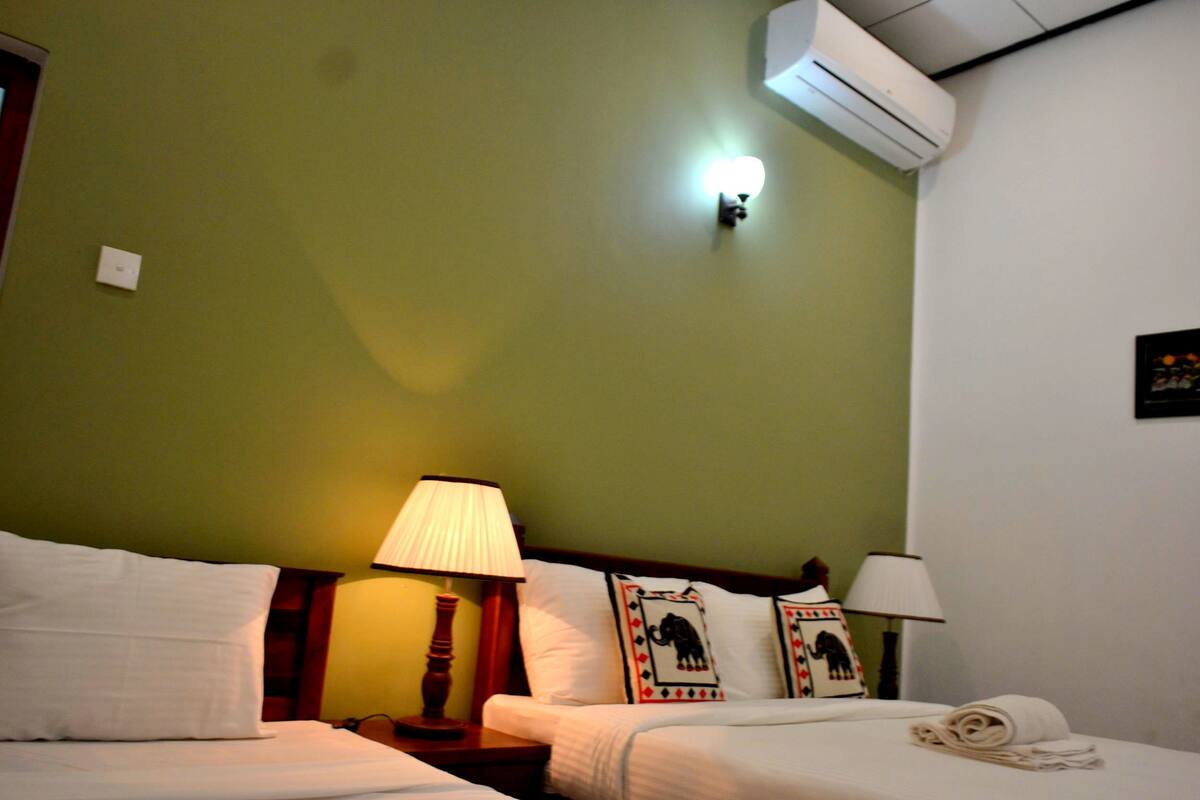 A hotel room with green walls, two beds, bedside lamps, a wall-mounted air conditioner, and decorative pillows.