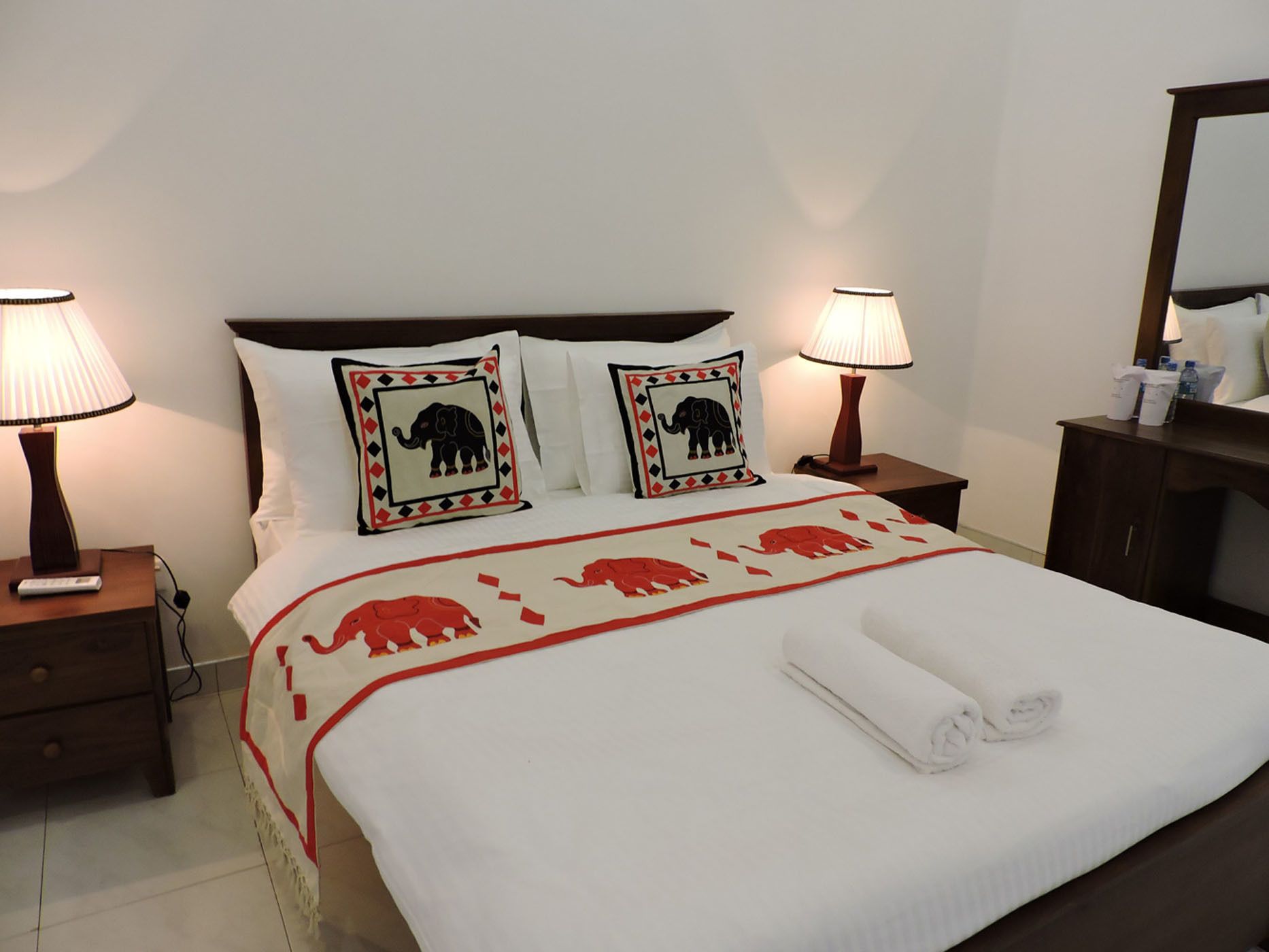 Una cama bien hecha con edredón blanco y dos almohadas con diseños de elefantes, flanqueada por dos mesitas de noche con lámparas.