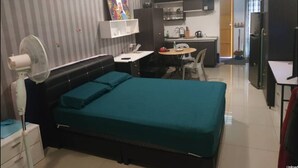 Una habitación de hotel compacta con cama, mesita de noche, ventilador y un área de kitchenette.