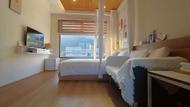 A modern bedroom with a bed, bedside table, TV, and a view of buildings through the window. - Tian Yan Mountains (Jiaoxi)