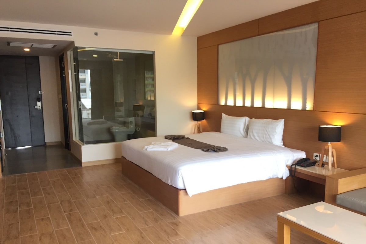 A modern hotel room with a large bed, bedside tables, a nightstand, a glass shower enclosure, and a wooden floor.