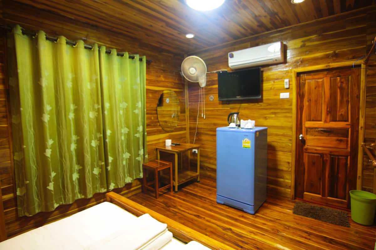 A wooden room with a bed, a small table, a blue refrigerator, a fan, and a television on the wall.