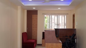 A reception area with a red chair, a wooden reception desk, a ceiling fan, and a window with blinds. - Sunnyvale Andaman (Port Blair)