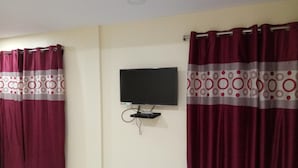 A room with a flat-screen TV mounted on the wall, a dark-colored sofa, and maroon curtains with a white and red pattern. - Sunnyvale Andaman (Port Blair)