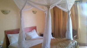 A bed with a canopy, a nightstand, and a chair in a room with beige walls and curtains. - Bondo Pride Hotel (Bondo)