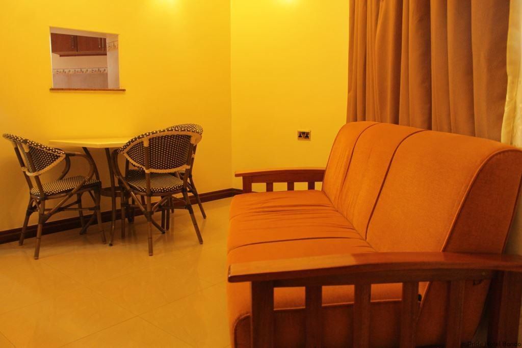 A room with a wooden sofa, two wicker chairs, a small table, and a kitchen window.