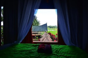 Una cama con ropa de cama verde y vistas a una construcción con techo de paja a través de cortinas translúcidas.