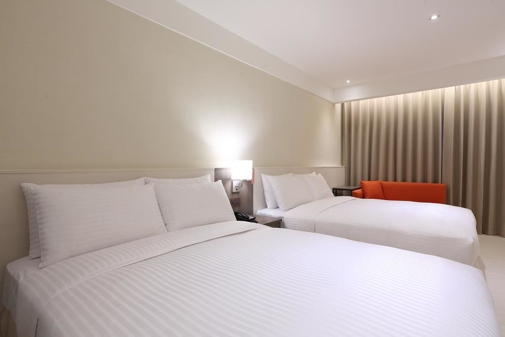 A hotel room with two beds, a nightstand, a lamp, and a curtain.
