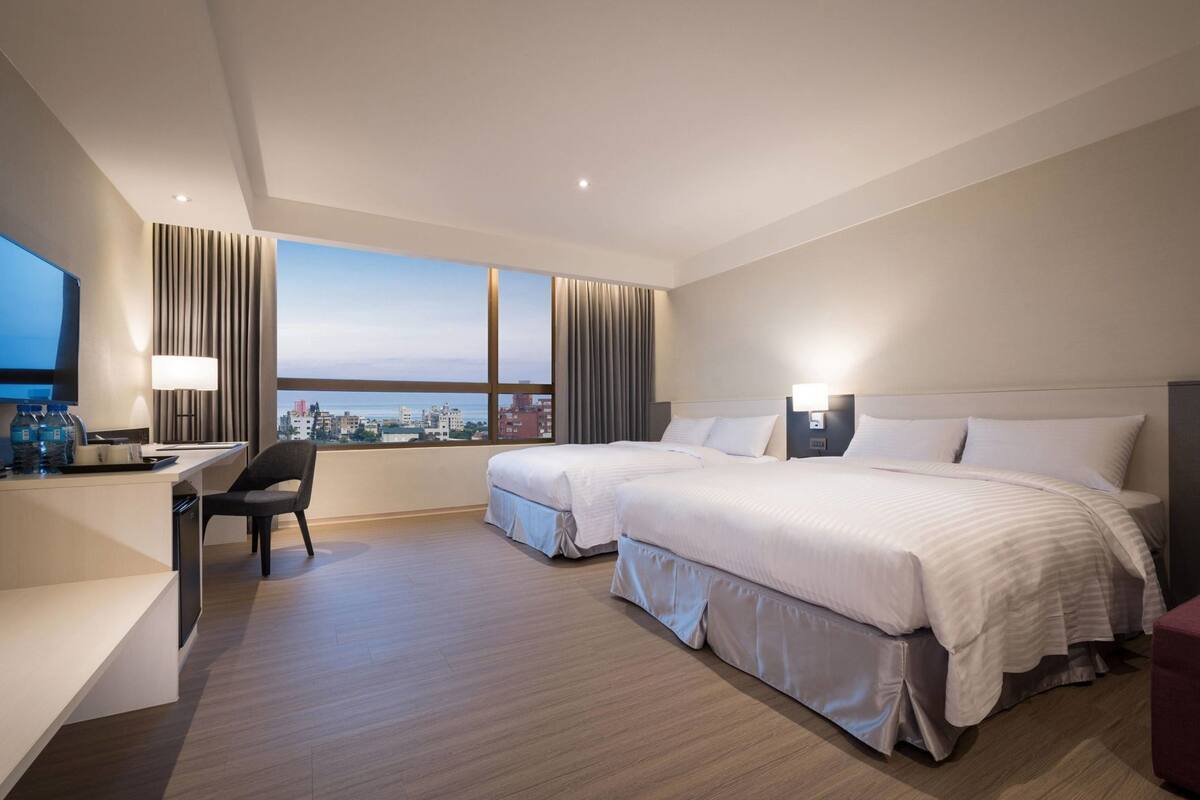Ein Hotelzimmer mit zwei Betten, einem Schreibtisch, einem Stuhl und einem großen Fenster mit Blick auf die Stadt.