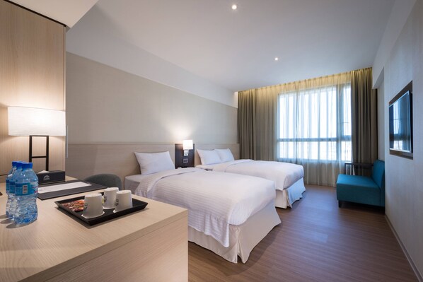 A hotel room with two beds, a desk with a tray of water bottles and cups, a TV, and a large window with curtains.