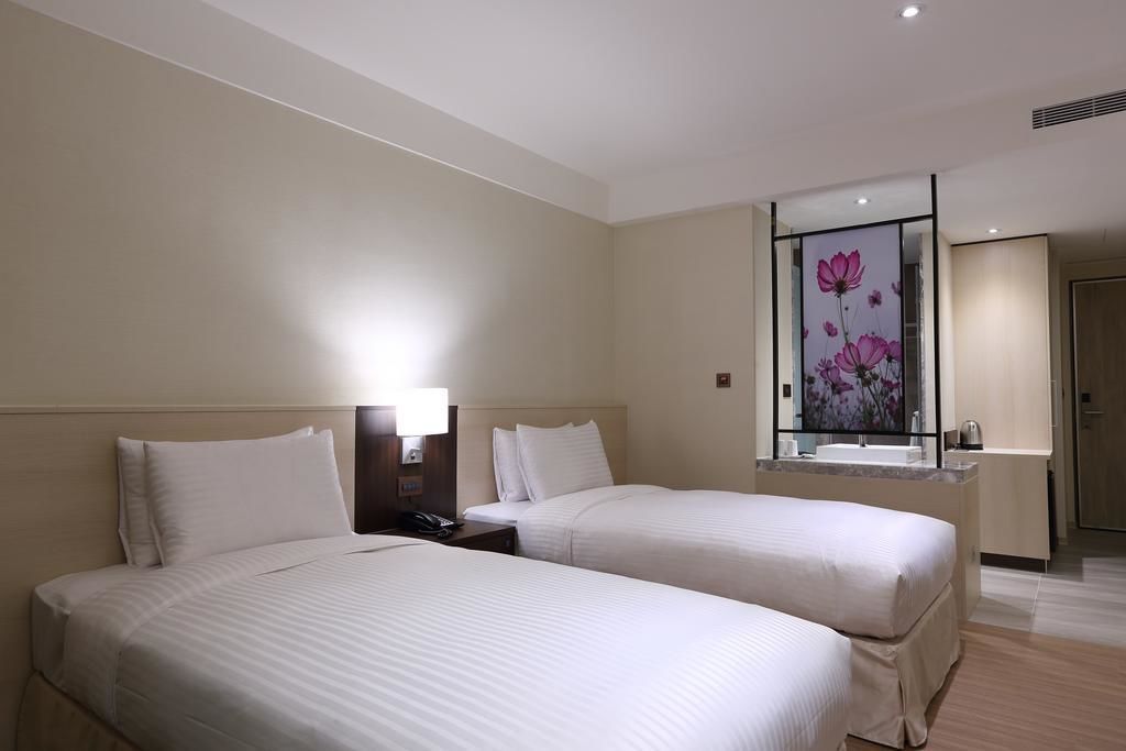 Habitación de hotel con dos camas, una mesita de noche, una lámpara, un espejo con diseño floral y un baño con lavabo y espejo.