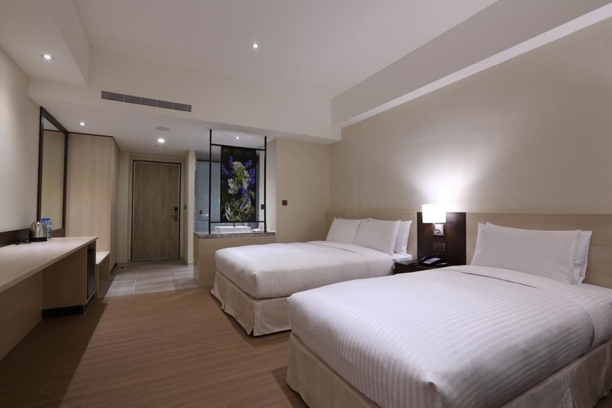 A hotel room with two beds, a desk, a mirror, and a window with a view.