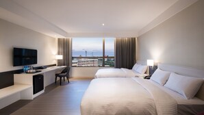 Habitación de hotel con dos camas, un escritorio con silla, televisor y vista a edificios y un cuerpo de agua.