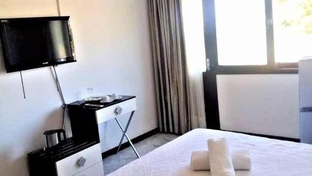 A hotel room with a bed, a desk with a flat-screen TV, a window with curtains, and a wall-mounted air conditioner. - Hotel Bilene (Praia do Bilene)