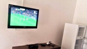 A room with a TV mounted on the wall displaying a sports event, a wooden cabinet with drawers, and a white refrigerator. - Hotel Bilene (Praia do Bilene)
