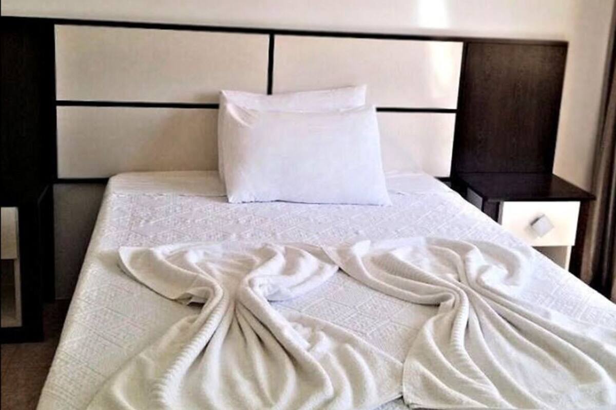 A neatly made bed with a white headboard and pillows, a bedside table, and a wall with a light switch.