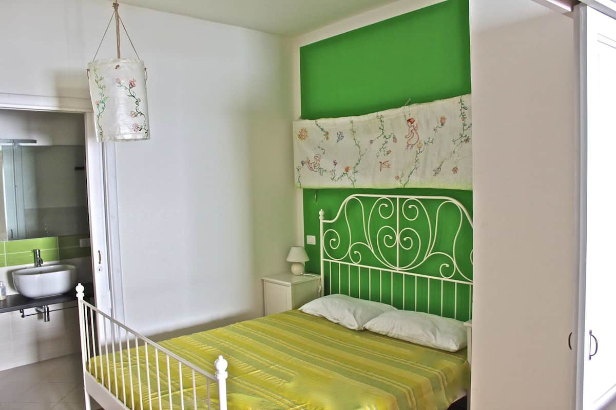 Un dormitorio con cabecera verde, cama con colcha amarilla a rayas, mesita de noche blanca con una lámpara, y una lámpara colgante.