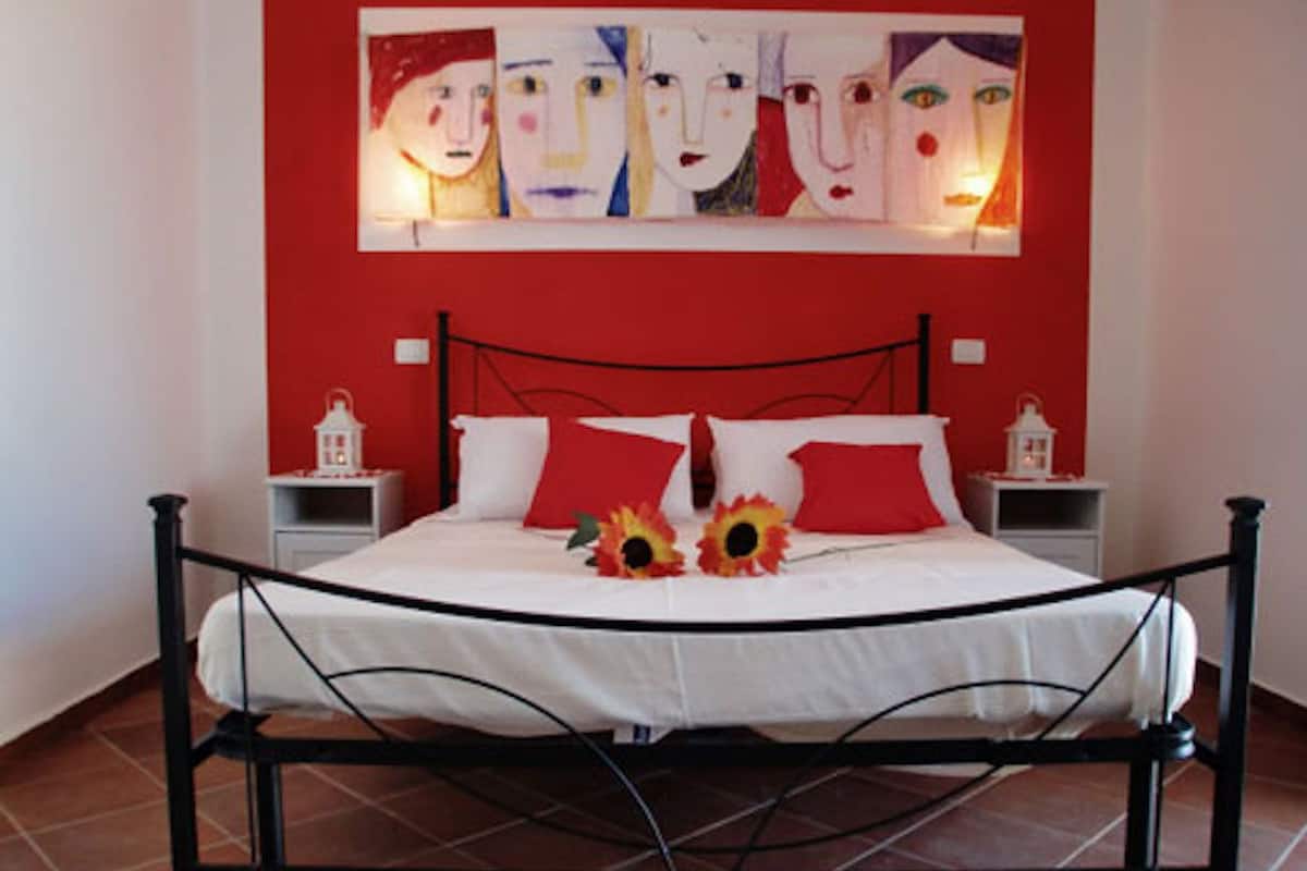 Una cama con cubrecama blanco, almohadas rojas, estructura metálica negra y dos mesitas de noche con faroles blancos.