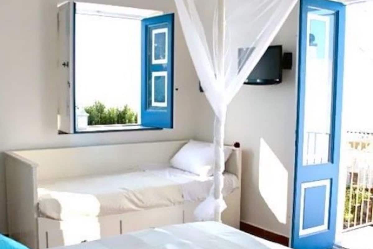 Un dormitorio con cama, ventana con contraventanas azules y puerta al balcón.
