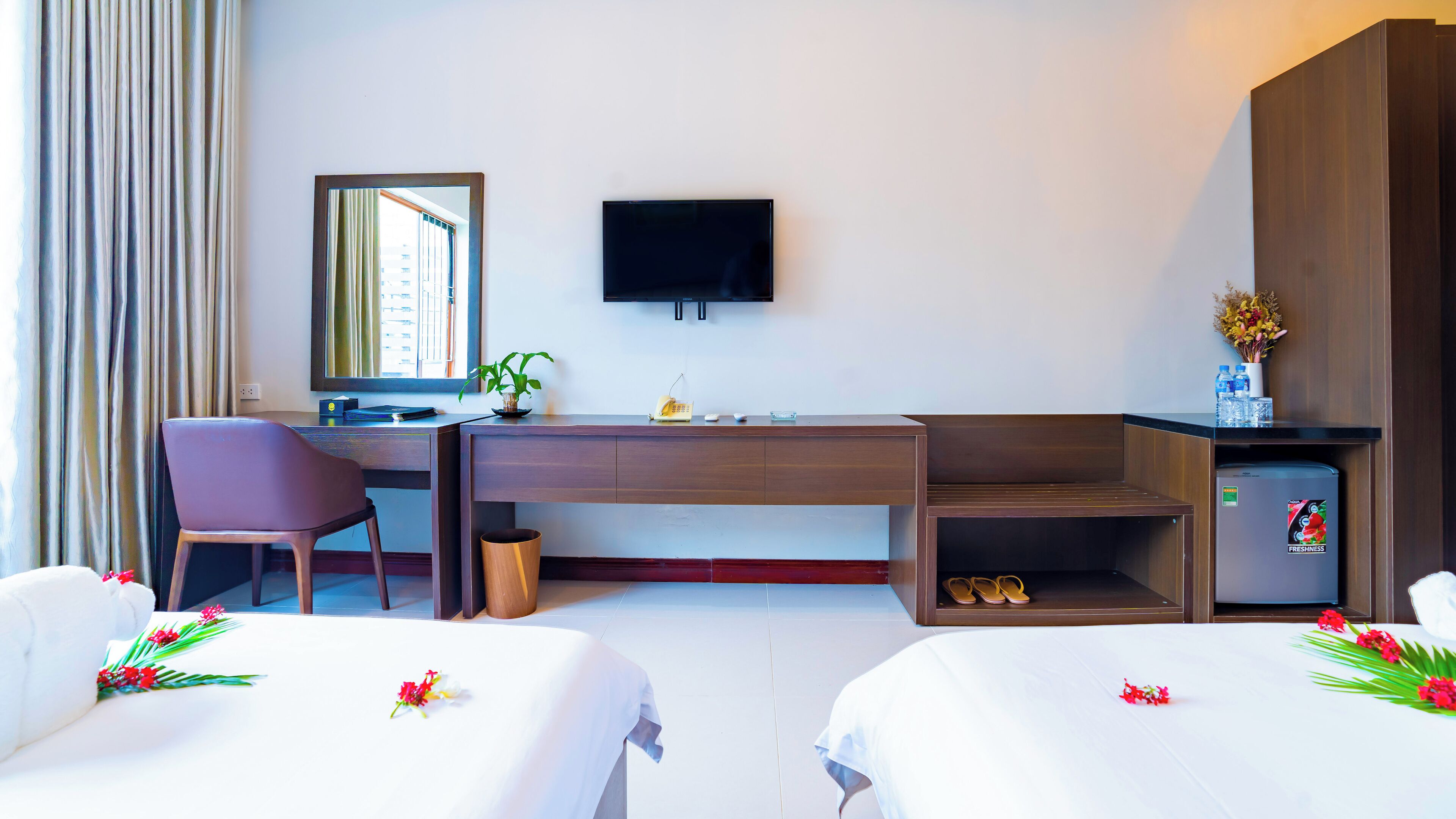 A hotel room with a desk, chair, two beds, a TV, a small fridge, and a window with curtains.