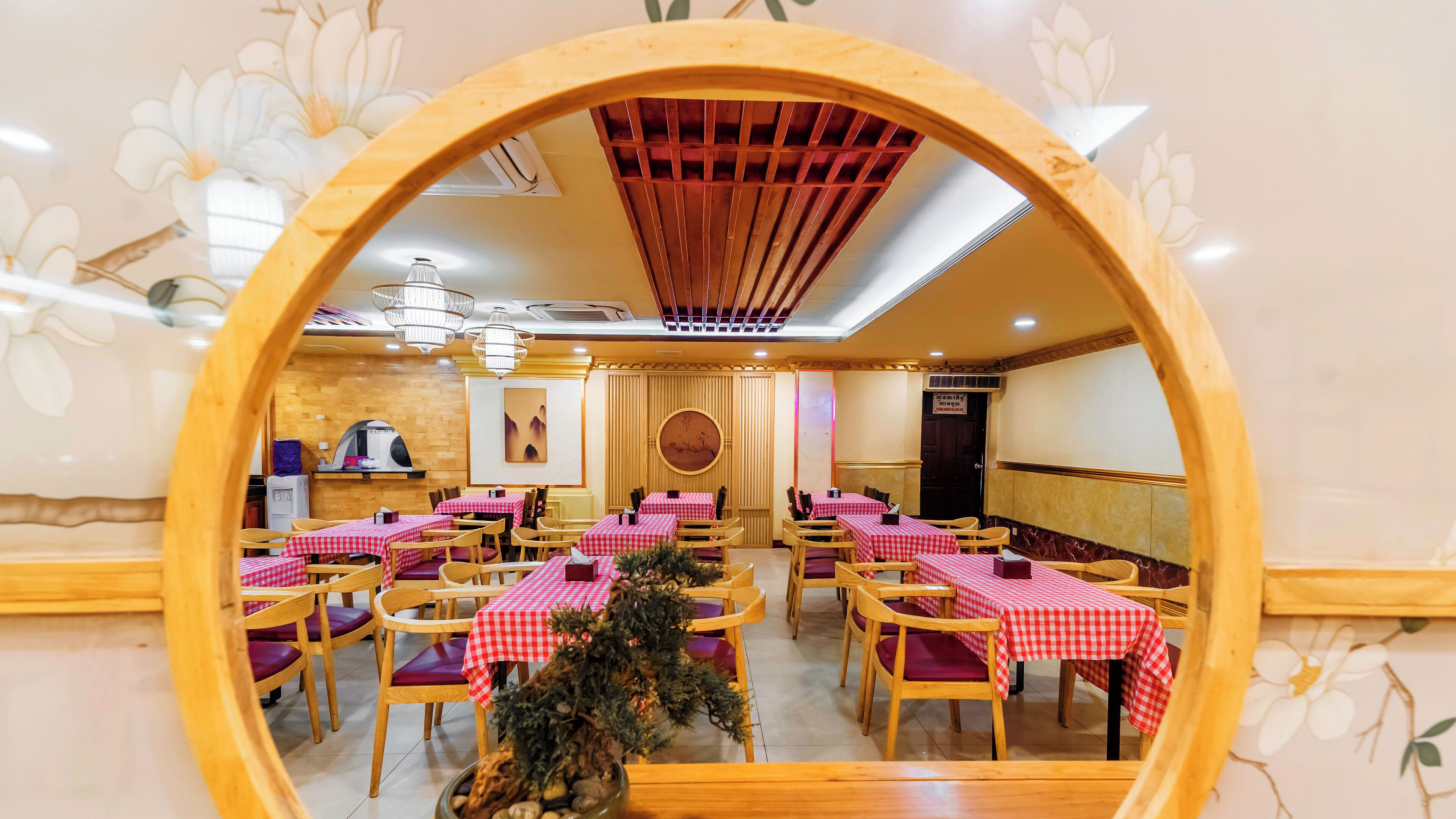 Restaurante com mesas redondas cobertas por toalhas xadrez vermelhas e brancas, cadeiras de madeira e um elemento decorativo central.