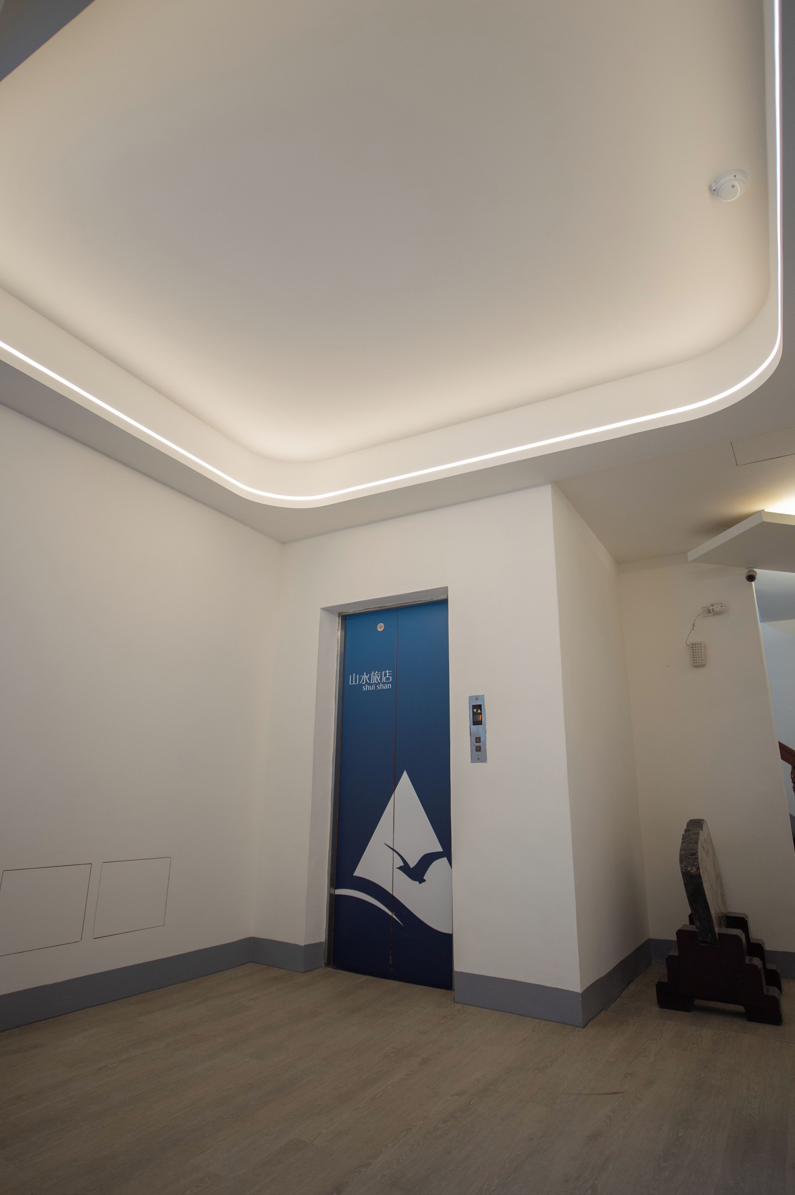 A modern elevator with a blue door and a logo, a plain wall, and a wooden floor.