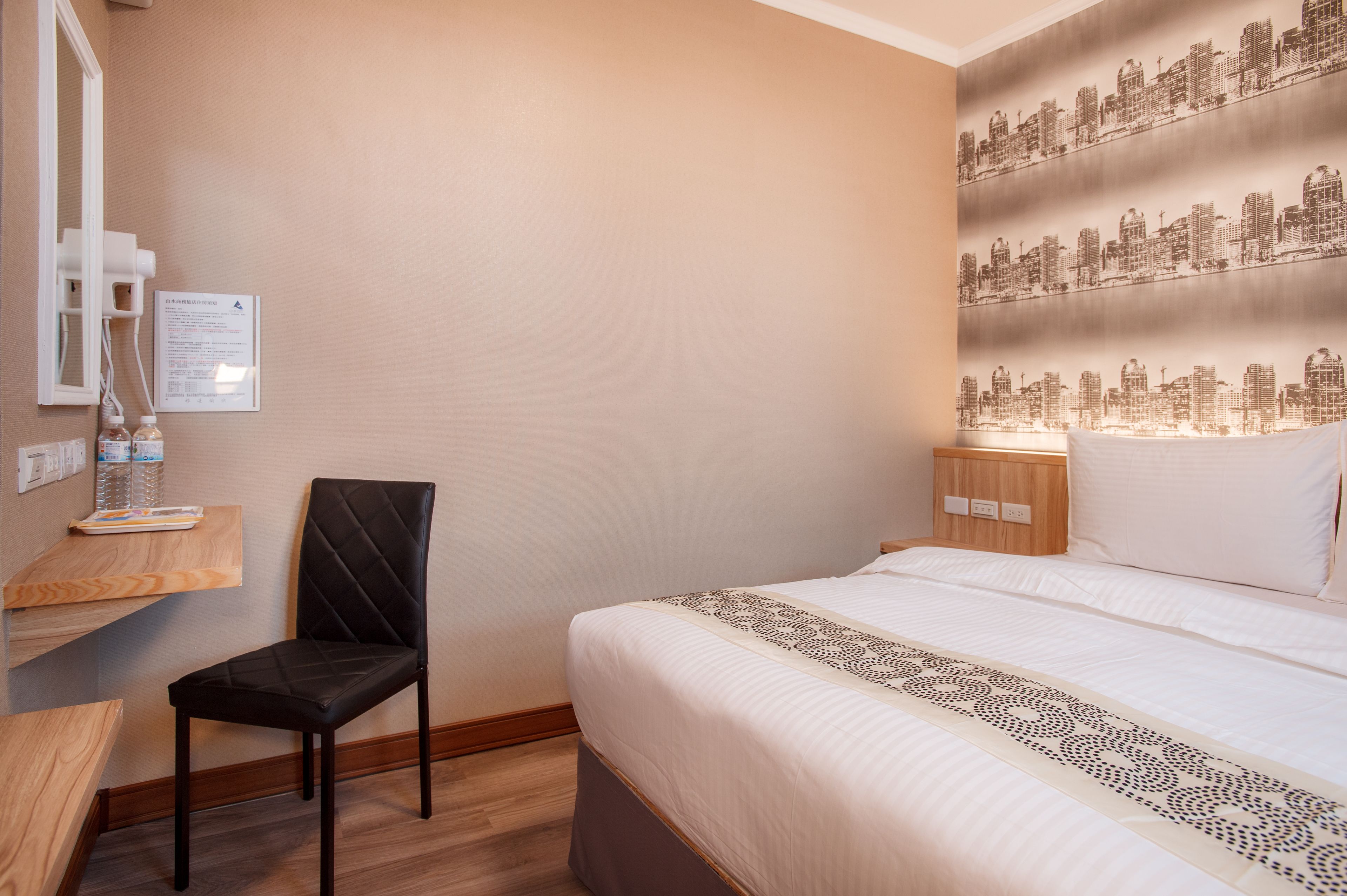 A hotel room with a bed, a chair, a nightstand, a wall-mounted mirror, and a cityscape wallpaper.